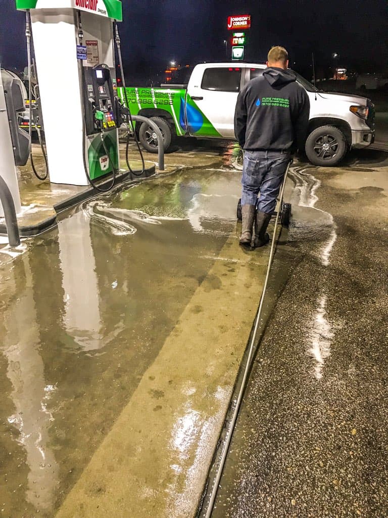 Worker using a pressure washer to clean wet pavement at a gas station at night.